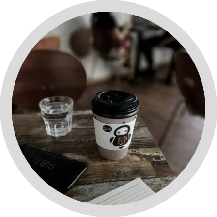 A CoffeeBot takeaway cup on a worn wooden table beside a glass of water and a notebook.